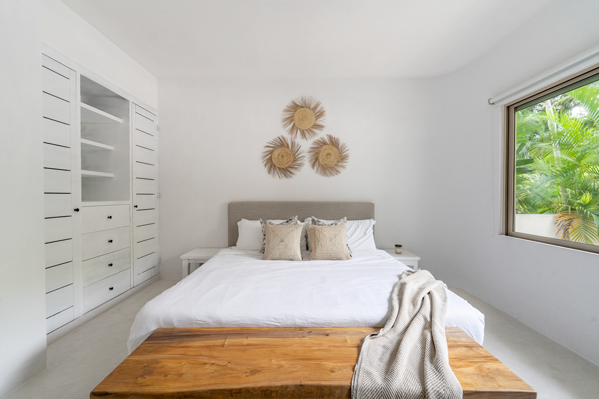 Sunlit bedroom with a wooden bed frame and white bedding — Real Zama